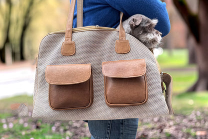 Beige pet carrier with brown leather pockets held by a person in an outdoor setting