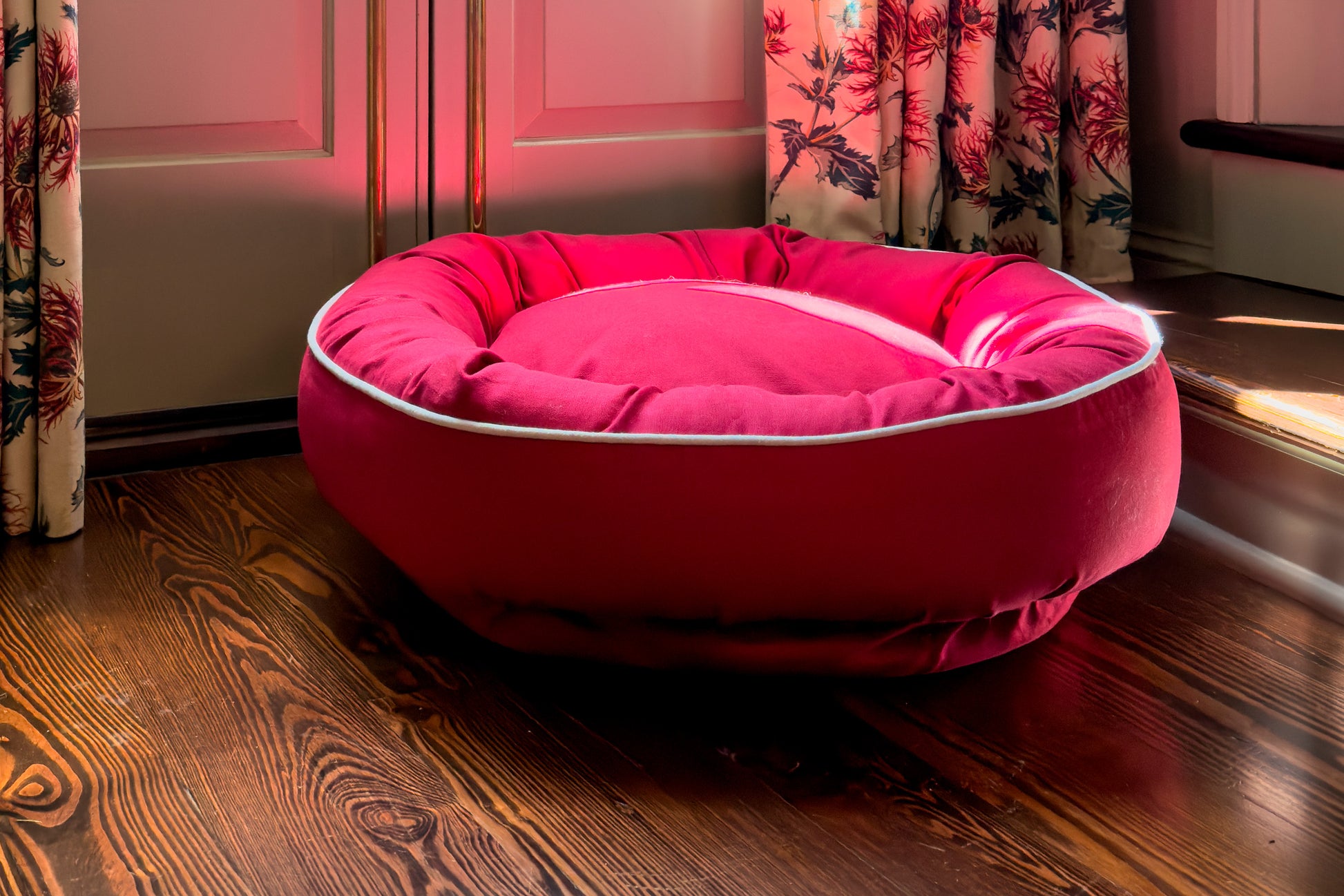 Red dog bed on a wooden floor with floral curtains in the background