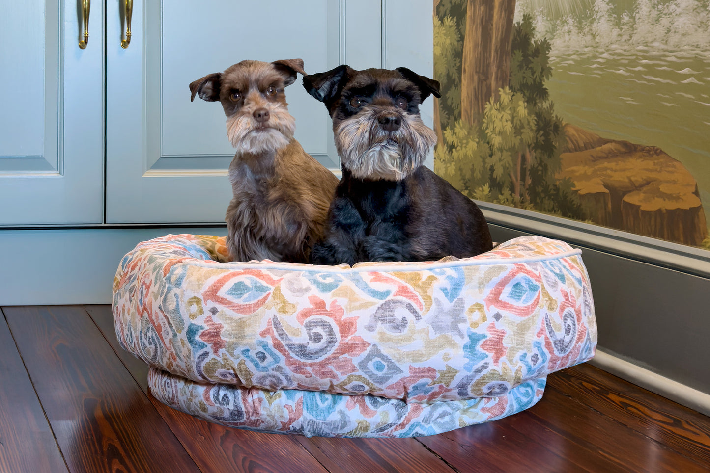 Two dogs sitting on a colorful patterned dog bed in a room with a painting on the wall.