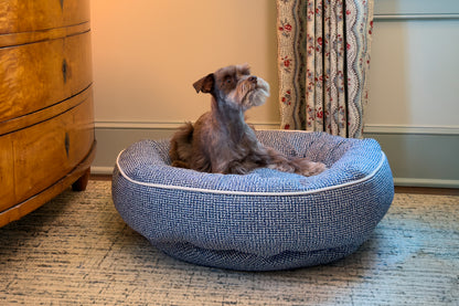 Dog lying on a dark denim dog bed in a room with a wooden dresser and floral curtain.
